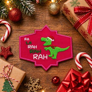 May include: A festive patch with a green dinosaur wearing a Santa hat and scarf, embroidered on a pink background. The patch reads "Fa La Rah Rah Rah" in white lettering. Surrounded by Christmas decorations, including candy canes and wrapped gifts.