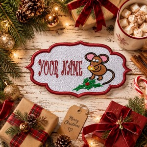 May include: A white embroidered patch with a red scalloped border featuring the text YOUR NAME in red, a brown mouse holding a yellow piece of cheese, and a green holly sprig with red berries, all set against a festive holiday background.