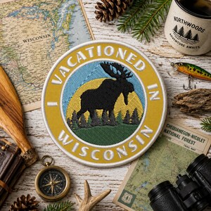 May include: A circular embroidered patch featuring a black moose silhouette against a yellow sun and green forest background. The border is mustard yellow with white embroidered text that reads I VACATIONED IN WISCONSIN.