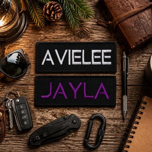 May include: Two rectangular black fabric patches with embroidered text. The top patch features the name AVIELEE in white capital letters, while the bottom patch displays the name JAYLA in purple capital letters with a clean, sans-serif font style.
