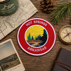 May include: A circular embroidered patch featuring a red border with the white text HOT SPRINGS ARKANSAS. The centre depicts a yellow sunburst over a blue lake, a wooden dock, and green pine trees against a dark silhouette of a forest.