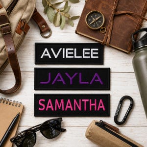 May include: Three rectangular black embroidered patches featuring the names AVIELEE in white, JAYLA in purple, and SAMANTHA in pink. The patches are arranged vertically on a white wooden surface surrounded by a backpack, notebook, compass, and sunglasses.