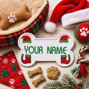 May include: A white bone-shaped patch with green embroidered text reading YOUR NAME, accented by four small embroidered elf hats and shoes in red and green. The patch is surrounded by a pet bed, a Santa hat, a ball, a rope toy, and treats.