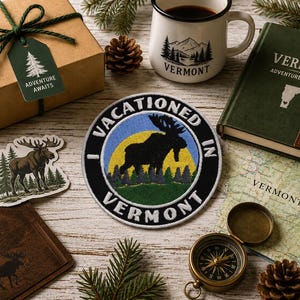 May include: A circular embroidered patch featuring a black moose silhouette against a yellow sun and blue sky, with the text I VACATIONED IN VERMONT. The patch is surrounded by a gift box, a Vermont journal, a mug, a compass, and a map.