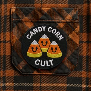 Może przedstawiać: Zbliżenie na brązowo-czarną koszulę w kratę z kieszenią. Kieszeń posiada okrągłą naszywkę z napisem "CANDY CORN CULT" wokół grafiki trzech uśmiechniętych cukierków kukurydzianych.