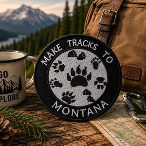 May include: A circular embroidered patch featuring a black border and a white centre. The patch displays black bear paw prints and the text "MAKE TRACKS TO MONTANA" in white capital letters. The design is centred on a dark fabric background.