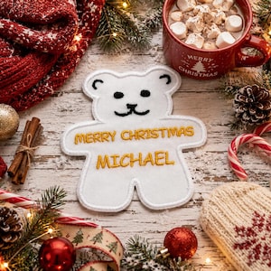 May include: A white felt bear-shaped ornament featuring black embroidered eyes and a mouth, with the gold embroidered text "MERRY CHRISTMAS" and "MICHAEL" centered on the chest, finished with a white stitched border edge.