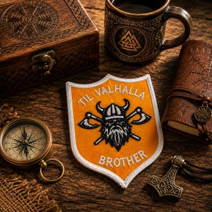 May include: A shield-shaped embroidered patch featuring an orange background with a white border. The patch displays a black and white graphic of a bearded warrior wearing a horned helmet, flanked by two crossed axes, with the text TIL VALHALLA BROTHER.