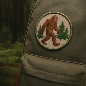 Pode incluir: Um patch redondo com um Bigfoot castanho a andar em frente a duas árvores de pinheiro verdes. O patch é cosido numa mochila verde-oliva. O fundo é uma cena florestal desfocada.