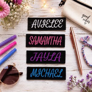 May include: Four rectangular black fabric patches with embroidered names in cursive script. The names are Avielee in white, Samantha in pink, Jayla in purple, and Michael in blue. Each patch features a clean, stitched border on a white wood surface.