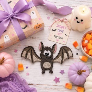 May include: An embroidered bat patch with brown fur, tan wings, and a white face, positioned near a gift box with a purple ribbon, a tag reading SPOOKY CUTE JUST FOR YOU!, a ghost-shaped candle, candy corn, and small pastel pumpkins.