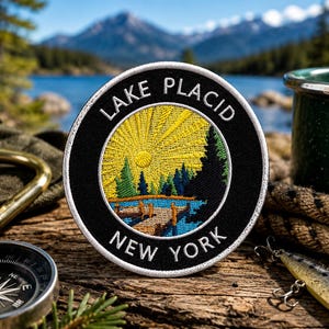 May include: Embroidered patch with the text "Lake Placid New York" in white on a black background. The patch features a yellow sunburst design over a landscape with trees, water, and a bridge. A compass, carabiner, and fishing lure are also visible.