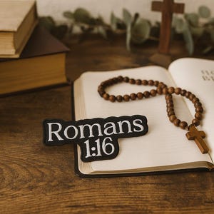 Puede incluir: Una Biblia abierta con un rosario y una cruz de madera sobre una superficie de madera. Un parche con el texto "Romanos 1:16" está sobre la Biblia. Una taza y libros apilados se encuentran en el fondo.