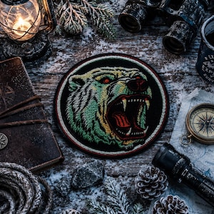 May include: A circular embroidered patch featuring a snarling polar bear head with red eyes, sharp teeth, and a black background. The bear is rendered in shades of white, yellow, and green thread, surrounded by a thin red border on the outer edge.