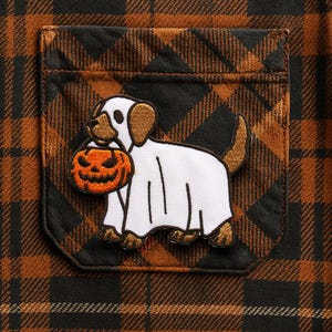 以下が含まれることがあります： 茶色と黒のチェック柄の生地のクローズアップ。ポケットには、オレンジ色のジャック・オー・ランタンを持った幽霊のコスチュームを着た犬の刺繍パッチが付いています。生地は質感のある外観をしています。
