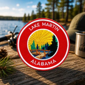 Peut inclure: Un écusson rond rouge Lake Martin Alabama avec du texte blanc et un motif brodé détaillé. Le motif représente un soleil jaune, des arbres verts et une scène de lac bleu. L'écusson est sur une surface en bois.