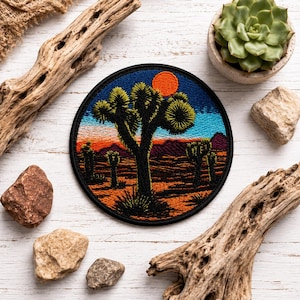 May include: A circular embroidered patch featuring a desert landscape with a prominent Joshua tree in the foreground, smaller cacti, and distant mountains under a vibrant sunset sky with a bright orange sun, all enclosed by a black stitched border.