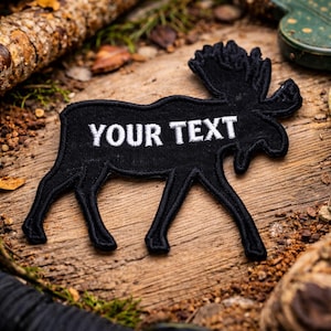 May include: A black embroidered patch in the shape of a moose, with the text "YOUR TEXT" in white. The patch is on a wooden surface with natural elements.