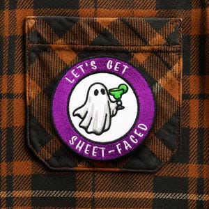 May include: Close-up of a brown and black plaid shirt pocket with a circular patch. The patch features a cartoon ghost holding a green cocktail, surrounded by the text "LET'S GET SHEET-FACED" in purple.