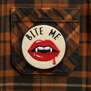 May include: Close-up of a brown and black plaid shirt pocket with a circular patch. The patch features red lips with fangs and the words "BITE ME" in black lettering. A drop of red liquid drips from the lips.