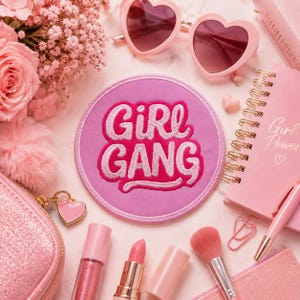 Op de afbeelding: Een ronde roze geborduurde patch met de tekst "GIRL GANG" in dikke, lichtroze letters. De patch is omringd door roze accessoires, waaronder een hartvormige zonnebril, een spiraalnotitieboek, een make-upkwast en een lippenstift.