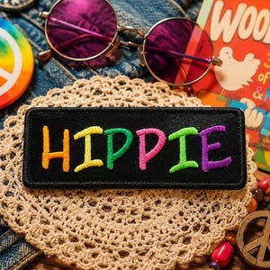 Könnte beinhalten: Eine Sammlung von Hippie-Artikeln, darunter ein schwarzer Aufnäher mit dem Wort "HIPPIE" in bunten Farben. Weitere Artikel sind runde Sonnenbrillen mit violetten Gläsern, ein Peace-Zeichen-Button und ein Vintage-"Woodstock"-Poster.