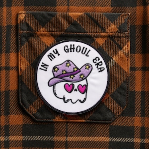 Puede incluir: Primer plano de un bolsillo de camisa a cuadros marrón y negro con un parche circular. El parche presenta una calavera de dibujos animados con un sombrero de vaquero morado con estrellas amarillas y ojos de corazón rosa. El texto "IN MY GHOUL ERA" está impreso alrededor de la calavera.