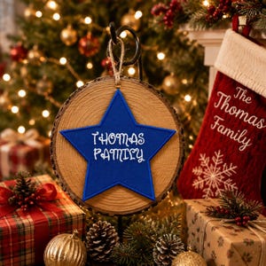 May include: A circular wood slice ornament featuring a blue felt star centered on the front. The star is embroidered with the white text "THOMAS FAMILY" in a decorative font. A jute twine loop is attached to the top for hanging purposes.