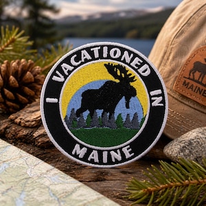 May include: A circular embroidered patch featuring a black moose silhouette against a yellow and light blue sky background, with green pine trees at the base. The black border contains the white embroidered text I VACATIONED IN MAINE.