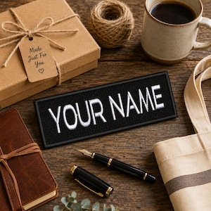 May include: A rectangular black embroidered patch featuring the white text "YOUR NAME" in a bold sans-serif font. The patch has a stitched border and is positioned on a wooden surface alongside a brown gift box, a pen, and a tote bag.