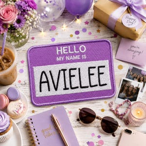 May include: A rectangular purple embroidered patch featuring the white text HELLO MY NAME IS above the name AVIELEE in black letters on a white background. The patch is surrounded by party decorations including flowers, a gift, and a notebook.