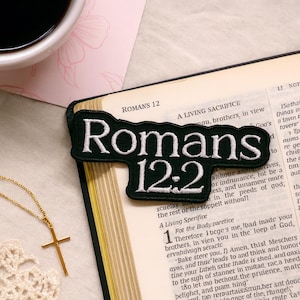 May include: A black embroidered patch with the text "Romans 12:2" in white, placed on an open book. A gold cross necklace and a cup of coffee are also visible.