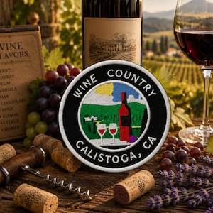 May include: A circular embroidered patch with a black border and white text reading WINE COUNTRY CALISTOGA, CA. The center features a colorful stitched landscape with a red wine bottle, two wine glasses, a yellow sun, and green vineyard rows.