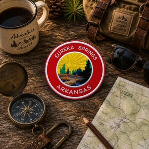 May include: A red and white embroidered patch with "Eureka Springs Arkansas" text and a landscape design. The image also includes a compass, map, coffee mug, backpack, and sunglasses, suggesting a travel theme.