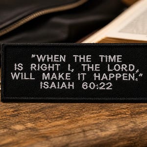 Pode incluir: Patch retangular preto com texto bordado branco: "QUANDO CHEGAR O TEMPO, EU, O SENHOR, FAREI ACONTECER", ISAÍAS 60:22. Uma cruz e uma corrente prateadas são visíveis. O patch está sobre uma superfície de madeira.