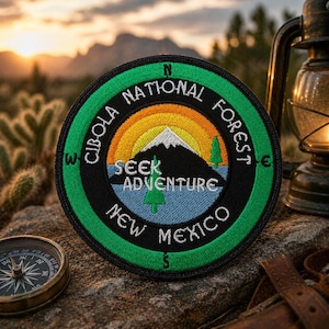 May include: Circular patch with a green border and the text "Cibola National Forest New Mexico." The patch features a mountain scene with a colorful sunset and the words "Seek Adventure."