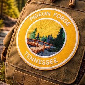 Puede incluir: Primer plano de una mochila marrón con un parche circular. El parche presenta un paisaje con un puente, árboles y un diseño de rayos de sol. El parche dice "PIGEON FORGE TENNESSEE" en letras blancas.