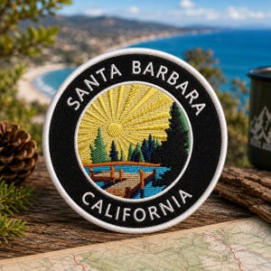 May include: A circular embroidered patch with the text "SANTA BARBARA CALIFORNIA" in white on a black background. The patch depicts a yellow sun, trees, and a bridge over water. The patch is on a wooden surface.