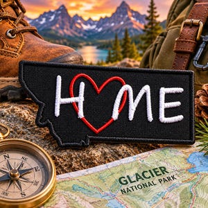 May include: A black patch with the word "HOME" in white, with a red heart replacing the "O", in the shape of the state of Montana. A compass, map, hiking boot, and backpack are in the background, suggesting a travel theme.