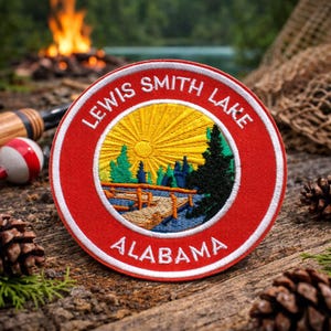 May include: A circular embroidered patch featuring a red border with white text reading LEWIS SMITH LAKE ALABAMA. The center depicts a yellow sun rising over a blue lake, a wooden dock, and green pine trees against a white background.