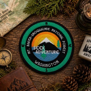 May include: A circular embroidered patch featuring a mountain landscape with a white peak, a blue base, and a yellow-to-orange sunset. The text reads Mt. Baker-Snoqualmie National Forest, Seek Adventure, and Washington, with compass directions.