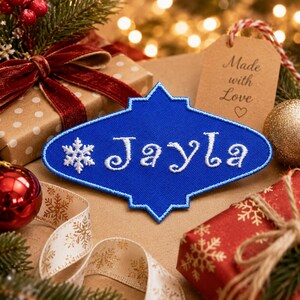 May include: A blue, quatrefoil-shaped embroidered patch featuring a white snowflake icon on the left and the name Jayla stitched in white cursive lettering. The patch has a blue stitched border and is displayed among holiday gift wrapping.