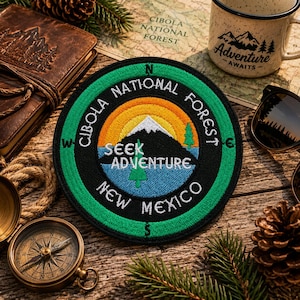 Puede incluir: Un parche bordado circular con un borde verde que muestra los puntos cardinales, un paisaje de montaña central bajo un sol amarillo y naranja, y el texto Cibola National Forest, New Mexico, y Seek Adventure en letras blancas.