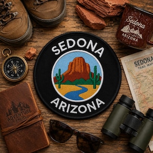 Può includere: Una toppa ricamata circolare raffigurante una formazione rocciosa rossa, due cactus saguaro verdi e un fiume blu su un cielo azzurro, con il testo bianco SEDONA ARIZONA arcuato lungo la parte superiore e inferiore del bordo nero.