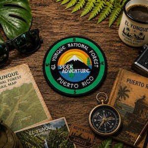May include: A circular embroidered patch featuring a mountain landscape with a sun, trees, and the text "EL YUNQUE NATIONAL FOREST PUERTO RICO" and "SEEK ADVENTURE" in the center. The patch has a black border and a green outer ring with compass points.