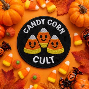 May include: A circular black embroidered patch featuring three smiling candy corn characters in orange, yellow, and white. The text CANDY CORN CULT is stitched in white thread around the top and bottom of the design on a black fabric background.