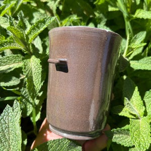 May include: A handmade brown ceramic mug with a small handle. The mug has a slightly textured surface and a simple, elegant design. The mug is surrounded by green mint leaves.