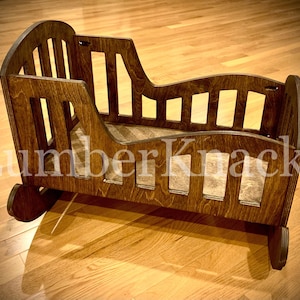May include: A wooden rocking cradle with a slatted design and a curved headboard. The cradle is made of dark brown wood and has a natural finish. The text "Lumber Knacks" is visible on the front of the cradle.