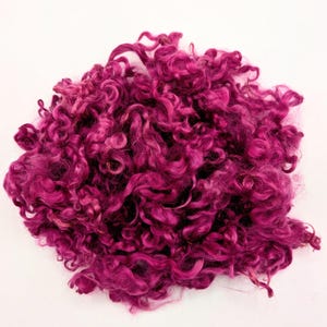 May include: A pile of vibrant, curly magenta-colored fiber, possibly wool or a similar material, against a white background. The fibers are densely packed, creating a textured, voluminous appearance. The color is consistent throughout, with no visible variations.
