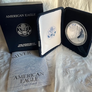 May include: A silver American Eagle one-ounce proof bullion coin in a presentation case. The coin is displayed in a black velvet case with a white interior. The box and paperwork are navy blue with silver text. The coin features an eagle design.